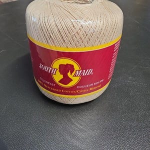 May include: A large ball of beige mercerized cotton thread, wrapped in clear plastic. The label is red with yellow accents and features the text "SOUTH MAID" and a silhouette of a woman's head. The thread is labeled as colorfast.