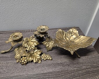 May include: A collection of antique brass candle holders and a leaf-shaped dish. The candle holders feature grape leaf and grape cluster designs. The leaf dish is a large, detailed leaf shape. The items are on a wooden surface.