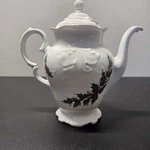 May include: White porcelain teapot with a lid, handle, and spout. The teapot features embossed floral designs and is decorated with holly and red berries. The rim of the lid and the top of the teapot have a gold trim.