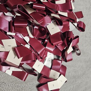 May include: A pile of burgundy satin ribbons with looped ends and small white tags. The ribbons are arranged in a dense, overlapping pattern, creating a textured surface. The tags have a small, simple design.