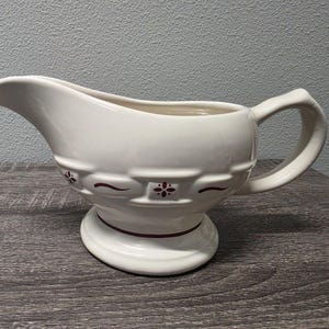 May include: A cream-colored ceramic gravy boat featuring a pedestal base and a curved handle. The exterior is decorated with a repeating embossed pattern of small maroon floral motifs and horizontal lines encircling the midsection of the vessel.