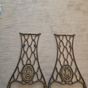 May include: Two antique Singer sewing machine legs. The legs are made of dark metal with an ornate, openwork design. Each leg features an oval emblem with the Singer logo. The legs are designed to support a sewing machine.