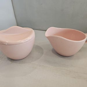 May include: A light pink, vintage sugar bowl with a lid and a matching creamer. The sugar bowl has a unique, curved lid design. The creamer has a handle and a spout. Both pieces are made of a smooth, solid material.