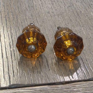 May include: Two amber-colored glass cabinet knobs with a faceted, octagonal shape. Each knob features a central circular metal insert with a textured, dark gray finish and a small metal screw protruding from the back for mounting purposes.