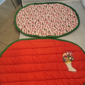 May include: Two oval Christmas placemats. One features a white background with red candy canes and green trim. The other is red with a quilted pattern, green trim, and a Christmas stocking applique.