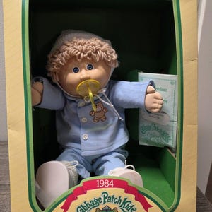 May include: A 1984 Cabbage Patch Kids doll with tan skin, blue eyes, and curly light brown yarn hair. The doll wears a light blue pajama set with a teddy bear graphic, a matching cap, and a yellow pacifier. It is inside a yellow cardboard box.