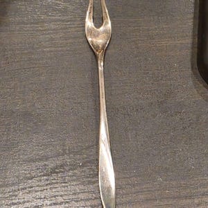 May include: A silver-toned metal cocktail fork featuring a slender, elongated handle and a two-pronged head. The utensil has a polished, reflective surface with a smooth, tapered design that narrows toward the base and flares slightly at the top.