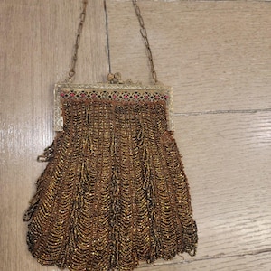 May include: An antique beaded handbag with a gold-colored metal frame and chain handle. The bag is covered in small, shimmering beads in shades of brown and gold, with a decorative top featuring red accents.