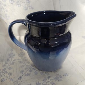 May include: A ceramic pitcher with a dark blue to light blue gradient glaze. The pitcher has a rounded body, a curved handle, and a spout for pouring. The background is a white cloth with a blue embroidered pattern.