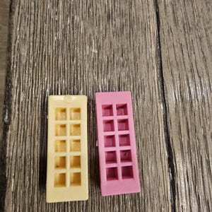 May include: Two miniature dollhouse windows. One is a pale yellow, the other is pink. Each window has a rectangular frame with a grid of small squares. The windows are made of plastic and are set against a wood grain background.