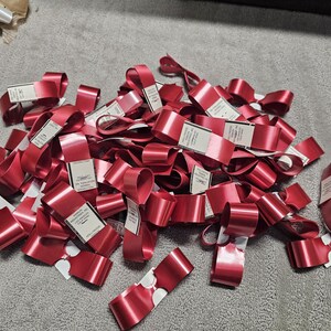 May include: A pile of shiny, red satin ribbons with white tags. The ribbons are looped and folded, creating a textured appearance. The tags have printed text. The ribbons are scattered on a gray surface.