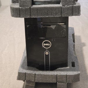 May include: A black Dell desktop computer tower secured in gray foam packaging. The front panel features a circular Dell logo, a power button, and a vertical vent design. The tower is positioned upright, showing the front face and top section.