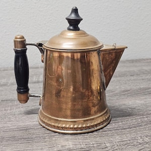 May include: A vintage copper coffee pot with a black wooden handle and lid finial. The pot has a spout and a hinged lid. The copper has a warm, aged patina, suggesting its antique nature. The handle is attached with a metal bracket.
