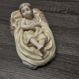 May include: A porcelain figurine of a baby lying in a manger. The baby has light skin and brown hair, wrapped in a cream-colored cloth. The manger is textured with a light brown finish on the sides and a smooth cream-colored interior base.