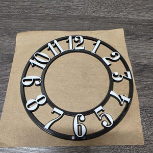 May include: A black clock face with white numerals from 1 to 12. The numbers are in a bold, sans-serif font. The clock face is circular and appears to be made of wood or a similar material. The clock face is on a brown paper.