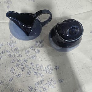 May include: A two-piece ceramic set in shades of blue. The set includes a small pitcher with a handle and a covered container. The pieces have a glossy finish and are placed on a white surface with a blue pattern.