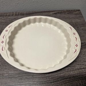 May include: A round, off-white ceramic pie dish with a fluted edge. The rim features a decorative pattern consisting of small, dark red floral motifs and curved lines embossed into the surface, providing a textured border around the smooth center.