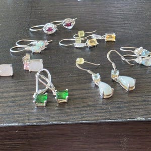 May include: A collection of various silver earrings featuring diverse gemstones, including pairs with green square-cut stones, teardrop-shaped white opalescent stones, pink rectangular stones, small red stones, and light blue marquise-cut stones.