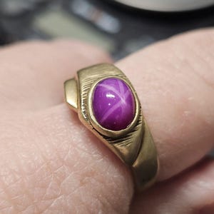May include: A gold-toned ring featuring a vibrant purple star ruby gemstone. The ring has a classic design with a rectangular setting and a smooth, polished band. The star ruby displays a six-rayed star pattern.