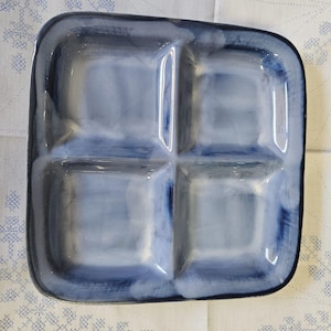May include: A square, divided serving dish with four compartments. The ceramic dish has a glossy finish and a watercolor-like design in shades of blue and white. The dish is set against a white background with a blue floral pattern.