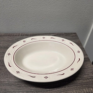 May include: An oval-shaped ceramic serving bowl in an off-white color. The rim features a decorative pattern consisting of small dark red floral motifs and curved lines, separated by a thin dark red circular border around the inner basin.