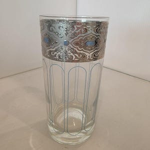 May include: A tall, clear glass with a silver filigree band at the top and light blue vertical lines. The silver band has a detailed, ornate design with small blue accents. The glass is suitable for serving beverages.