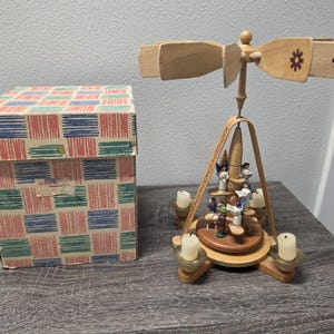 May include: A wooden Christmas pyramid with a spinning top and candle holders. The pyramid features small figurines and is accompanied by a patterned box. The spinning top has decorative cutouts.
