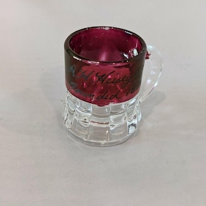 May include: A small, clear glass mug with a ruby red interior and rim. The mug has a clear handle and a base with a grid pattern. The text "A.H. Heisey, Founded 1896" is written on the side.