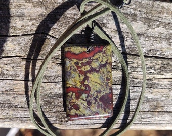 May include: A rectangular pendant featuring a mottled pattern of olive green, deep red, and dark brown tones. The pendant is suspended from a thin, olive green suede cord that is secured with dark metal coils at the top of the necklace loop.