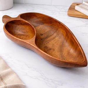 May include: A wooden serving bowl with a leaf-like design. The bowl is made of a warm-toned wood and features a large, elongated section and a smaller, attached bowl. Ideal for serving snacks or appetizers.
