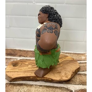 May include: A brown figurine of a Polynesian man with black curly hair, tattoos, and a green leaf skirt. The figurine is standing on a wooden cutting board with a hole. The background is a white tiled wall.