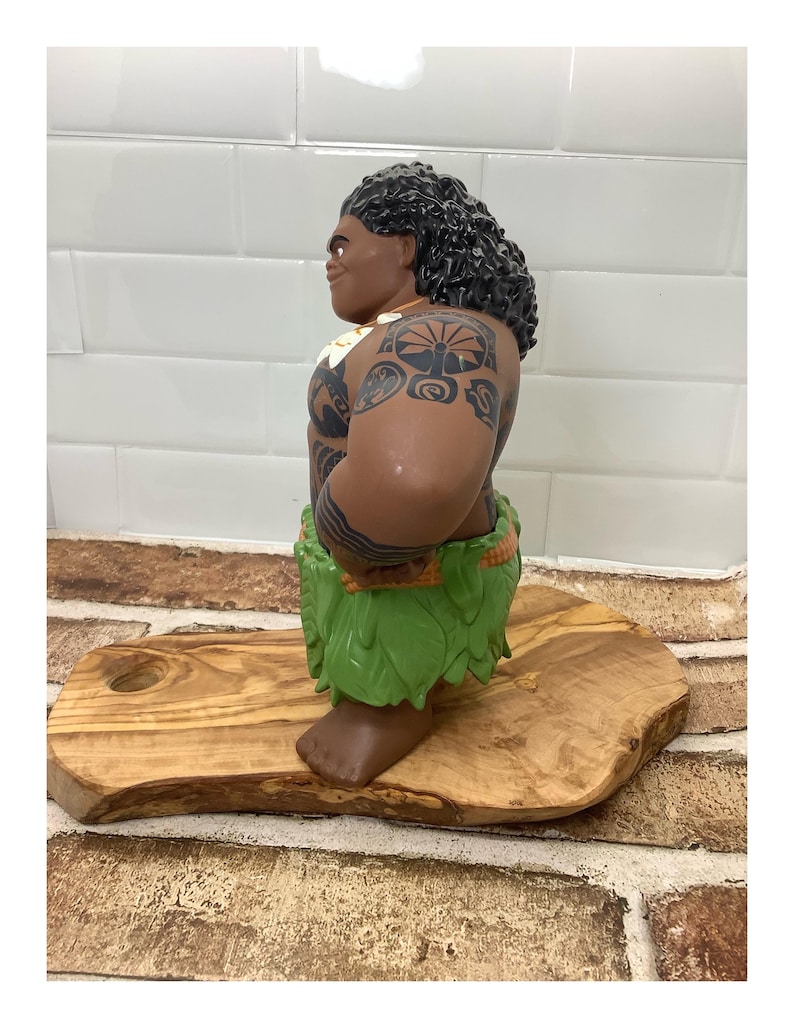 May include: A brown figurine of a Polynesian man with black curly hair, tattoos, and a green leaf skirt. The figurine is standing on a wooden cutting board with a hole. The background is a white tiled wall.