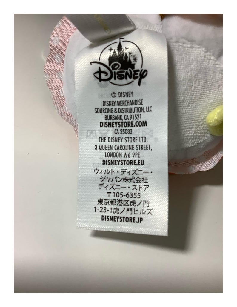 May include: A white tag with the Disney logo and text. The tag includes the Disney copyright information, the address for Disney Merchandise Sourcing & Distribution, and the address for The Disney Store Ltd.