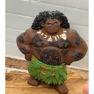 May include: A brown figurine of a muscular character with black curly hair, tattoos, and a green leaf skirt. The figurine has a necklace and is posed with hands on hips. The figure is standing on a wooden surface.