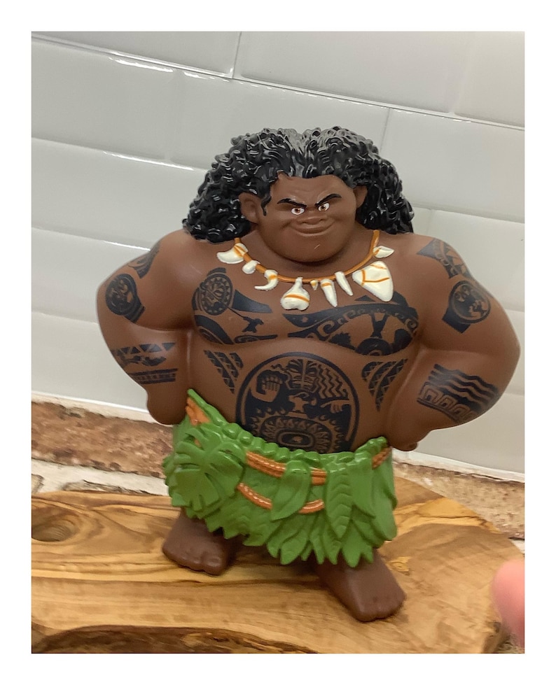 May include: A brown figurine of a muscular character with black curly hair, tattoos, and a green leaf skirt. The figurine has a necklace and is posed with hands on hips. The figure is standing on a wooden surface.
