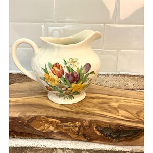 May include: Cream-colored ceramic pitcher with a floral design featuring tulips, daffodils, and other flowers. The pitcher has a curved handle and a scalloped rim. It sits on a wooden surface.