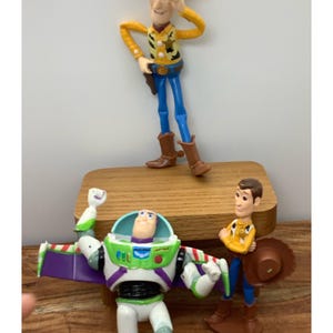 May include: A collection of Toy Story character figurines, including Woody in a cowboy hat and Buzz Lightyear with his wings extended. The figures are brightly coloured and stand on a wooden surface.