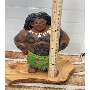 May include: A brown figurine of a Polynesian man with tattoos, wearing a green grass skirt and a necklace. The figurine is approximately 10 inches tall and is standing on a wooden base.