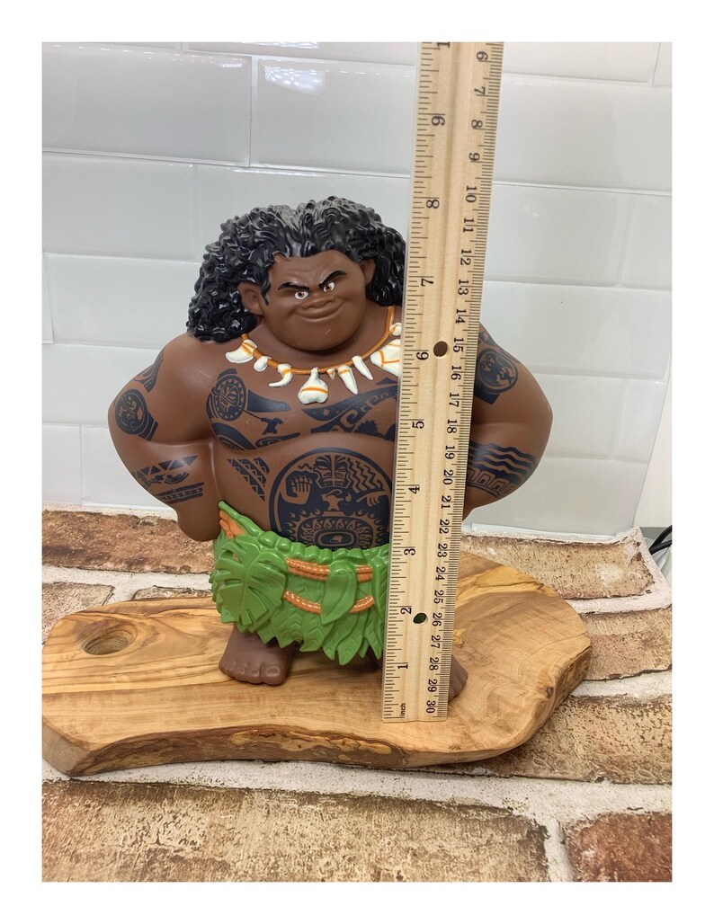 May include: A brown figurine of a Polynesian man with tattoos, wearing a green grass skirt and a necklace. The figurine is approximately 10 inches tall and is standing on a wooden base.