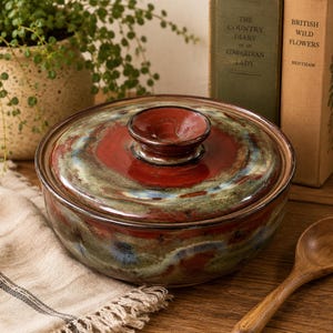 May include: A ceramic lidded casserole dish with a rich, swirled glaze in shades of red, green, and blue. The lid features a small, round handle. The dish is set on a wooden surface, with a wooden spoon and vintage books in the background.