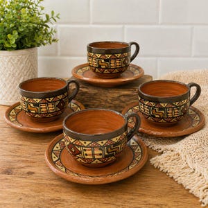 May include: A set of four terracotta-colored ceramic teacups and saucers. Each cup and saucer features a geometric pattern in black, yellow, and red. The cups have a dark brown rim and handle. The set is displayed on a wooden surface.