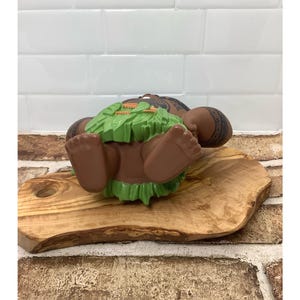 May include: A brown figurine of a Polynesian warrior with tattoos, lying on its back. The figurine has a green grass skirt and is resting on a wooden cutting board. The background features a white tiled wall and a brick wall.