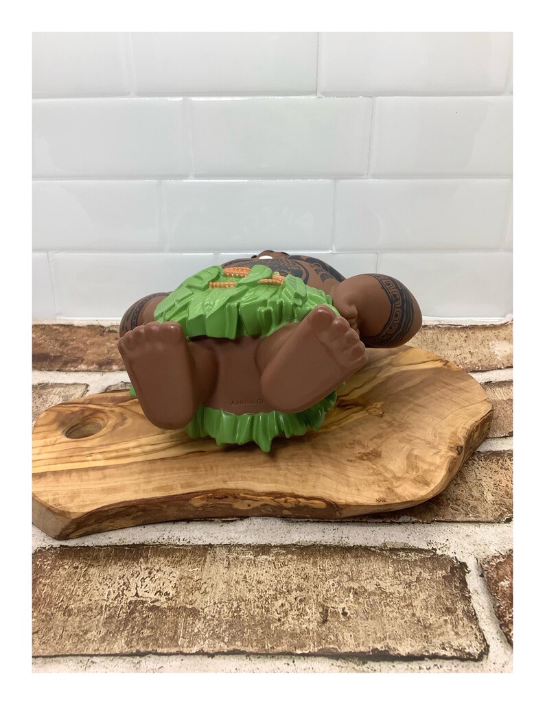 May include: A brown figurine of a Polynesian warrior with tattoos, lying on its back. The figurine has a green grass skirt and is resting on a wooden cutting board. The background features a white tiled wall and a brick wall.