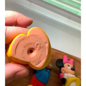Pu&ograve; includere: Primo piano di una statuetta Disney gialla e rossa, con interno rosa. La base della statuetta riporta la scritta "CHINA DISNEY" e altri marchi. &Egrave; visibile anche una statuetta di Minnie Mouse.