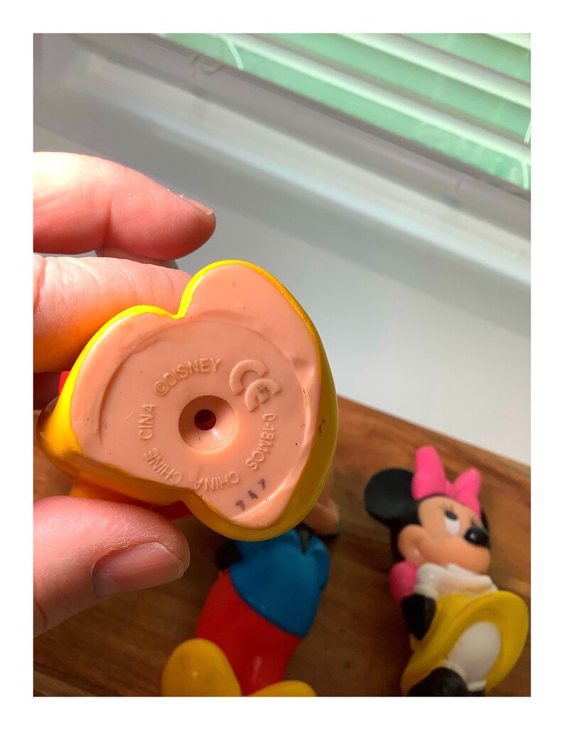 Pu&ograve; includere: Primo piano di una statuetta Disney gialla e rossa, con interno rosa. La base della statuetta riporta la scritta "CHINA DISNEY" e altri marchi. &Egrave; visibile anche una statuetta di Minnie Mouse.