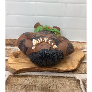 May include: A brown Polynesian-style figurine with black tattoos and a green leaf headdress. The figurine is lying on a wooden cutting board. The figurine has a shell necklace and black curly hair.