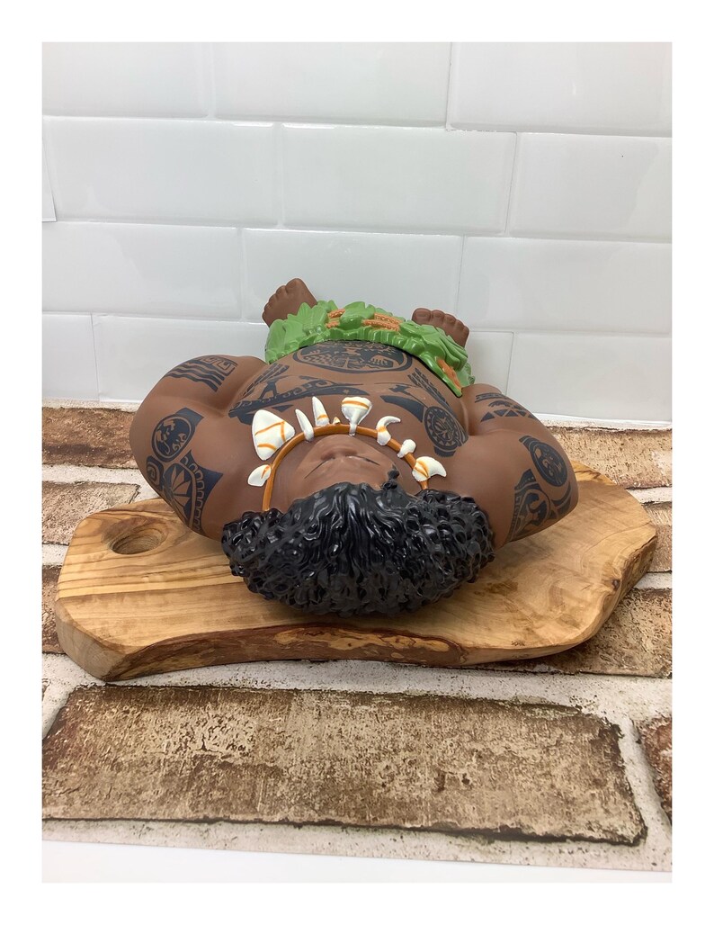 May include: A brown Polynesian-style figurine with black tattoos and a green leaf headdress. The figurine is lying on a wooden cutting board. The figurine has a shell necklace and black curly hair.