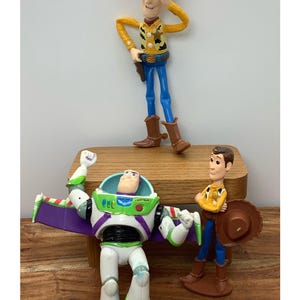 May include: Toy Story character figurines of Woody, Buzz Lightyear, and another Woody figure. Woody wears a cowboy hat, yellow shirt, blue trousers, and brown boots. Buzz Lightyear is in a white, green, and purple spacesuit. The other Woody figure is holding a hat.