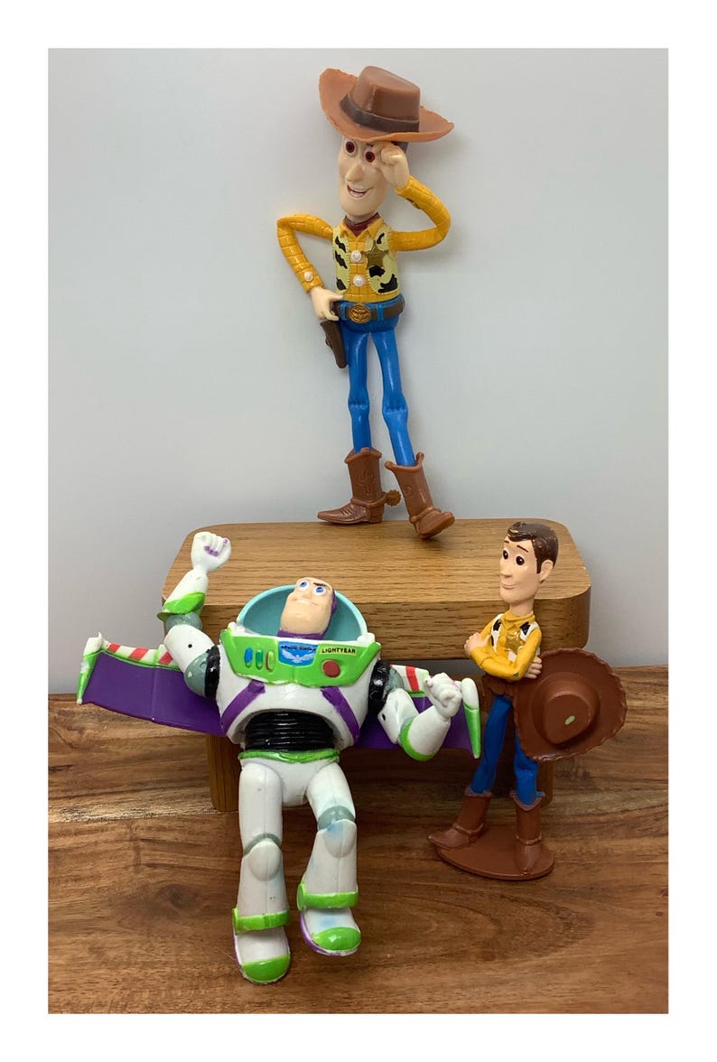 May include: Toy Story character figurines of Woody, Buzz Lightyear, and another Woody figure. Woody wears a cowboy hat, yellow shirt, blue trousers, and brown boots. Buzz Lightyear is in a white, green, and purple spacesuit. The other Woody figure is holding a hat.