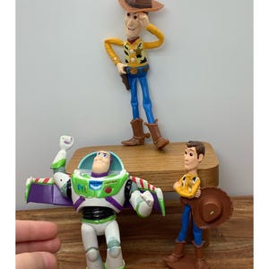 May include: Three Toy Story character figurines: Woody, Buzz Lightyear, and another Woody. Woody is wearing a cowboy hat, yellow shirt, blue trousers, and brown boots. Buzz Lightyear is in a white and green spacesuit. The other Woody is holding a hat.